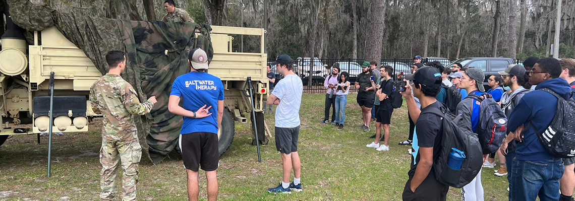 Capstone students watching members of the US Army 3rd infantry division perform a camouflage demonstration by concealing a military truck under a camouflage material.